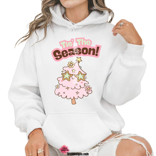 Christmas Sweaters by KrcilDesigns.com | Graphic design on a white hoodie, features a cute pink Christmas tree wearing star-shaped sunglasses, decorated with pastel stars and pastel daisy flowers, with text that reads: "Tis' The Season!" in bold retro-style pink, green, and brown lettering curved above the tree.