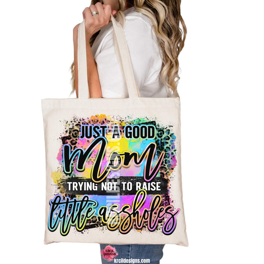 Sassy Mom Tote Bags by KrcilDesigns.com | Graphic design on a canvas tote bag featuring bold, colorful text that reads "Just A Good Mom Trying Not To Raise Little Assholes" The word "Mom" is in a vibrant rainbow script with leopard print accents, and the background includes a splash of bright paint textures in yellow, teal, pink, and purple. The style is playful, edgy, and sassy—perfect for fun-loving moms with a sense of humor.