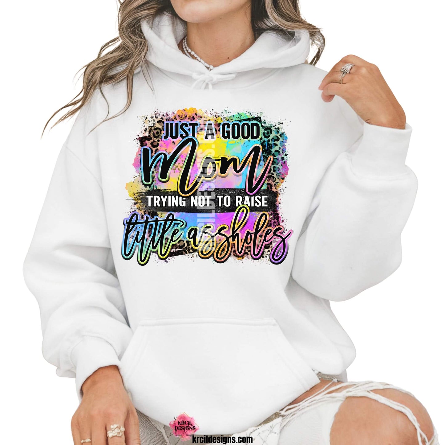 Sassy Mom Sweatshirts and Hoodies by KrcilDesigns.com | Graphic design on a white hoodie featuring bold, colorful text that reads "Just A Good Mom Trying Not To Raise Little Assholes" The word "Mom" is in a vibrant rainbow script with leopard print accents, and the background includes a splash of bright paint textures in yellow, teal, pink, and purple. The style is playful, edgy, and sassy—perfect for fun-loving moms with a sense of humor.