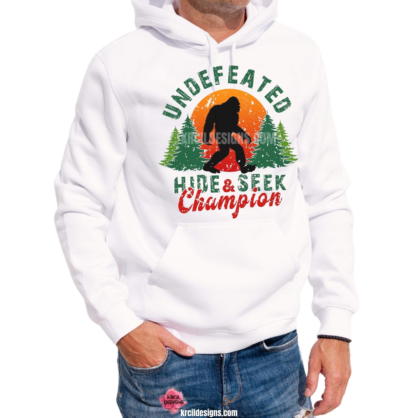 Bigfoot Shirts by KrcilDesigns.com | Graphic design on a white hoodie, featuring a graphic of a black silhouette of Bigfoot, walking in front of a red and orange sunset, with green pine trees on both sides, above Bigfoot, text that reads: "UNDEFEATED" in curved distressed bold block capital green letters, below, text that reads: "HIDE & SEEK" in distressed bold block capital green letters, with "Champion" overlay in bold red handwriting script.