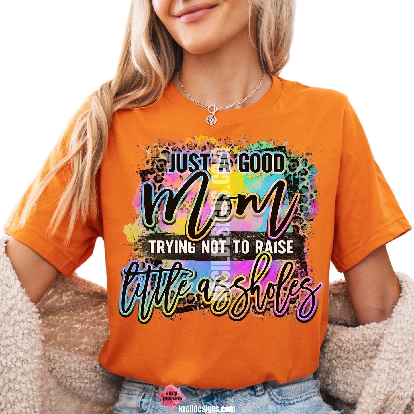 Sassy Mom Graphic Tees T-Shirts by KrcilDesigns.com | Graphic design on a orange t-shirt featuring bold, colorful text that reads "Just A Good Mom Trying Not To Raise Little Assholes" The word "Mom" is in a vibrant rainbow script with leopard print accents, and the background includes a splash of bright paint textures in yellow, teal, pink, and purple. The style is playful, edgy, and sassy—perfect for fun-loving moms with a sense of humor.