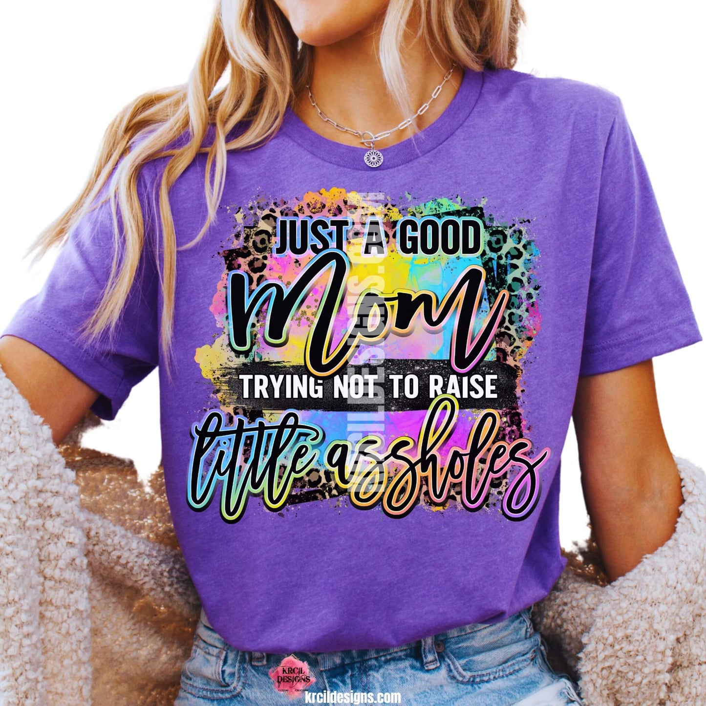 Sassy Mom Graphic Tees T-Shirts by KrcilDesigns.com | Graphic design on a purple t-shirt featuring bold, colorful text that reads "Just A Good Mom Trying Not To Raise Little Assholes" The word "Mom" is in a vibrant rainbow script with leopard print accents, and the background includes a splash of bright paint textures in yellow, teal, pink, and purple. The style is playful, edgy, and sassy—perfect for fun-loving moms with a sense of humor.