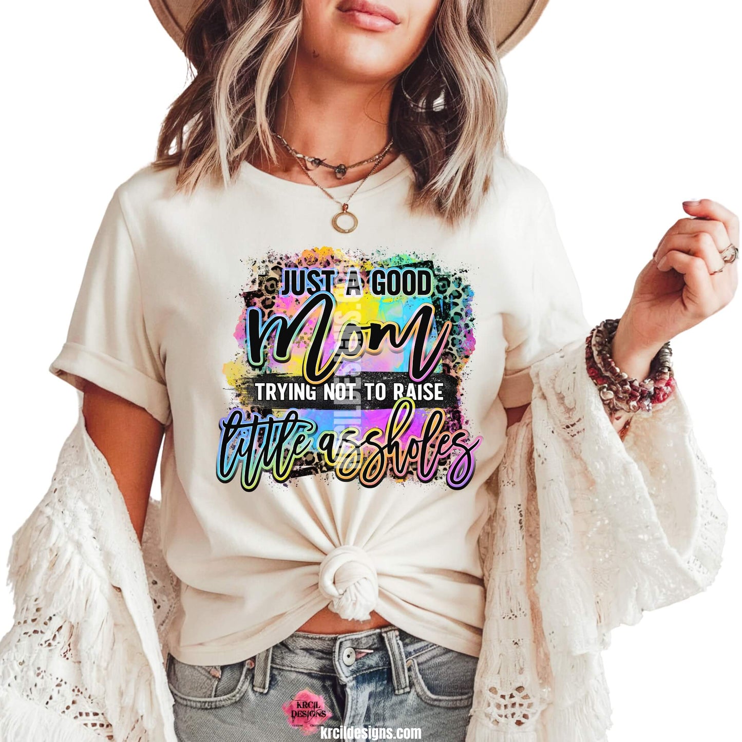 Sassy Mom Graphic Tees T-Shirts by KrcilDesigns.com | Graphic design on a white t-shirt featuring bold, colorful text that reads "Just A Good Mom Trying Not To Raise Little Assholes" The word "Mom" is in a vibrant rainbow script with leopard print accents, and the background includes a splash of bright paint textures in yellow, teal, pink, and purple. The style is playful, edgy, and sassy—perfect for fun-loving moms with a sense of humor.