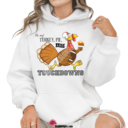 Funny Thanksgiving Shirts by KrcilDesigns.com | Graphic design on a white hoodie, featuring a cartoon turkey wearing a football helmet, holding a slice of pie topped with a dollop of whip cream on a white dinner plate, holding a football tucked under arm, and text that reads: "Oh, my! Turkey, Pie, and Touchdowns" in playful different fonts.