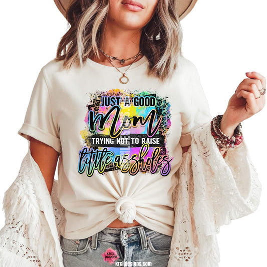 Sassy Mom Graphic Tees T-Shirts by KrcilDesigns.com | Graphic design on a white t-shirt featuring bold, colorful text that reads "Just A Good Mom Trying Not To Raise Little Assholes" The word "Mom" is in a vibrant rainbow script with leopard print accents, and the background includes a splash of bright paint textures in yellow, teal, pink, and purple. The style is playful, edgy, and sassy—perfect for fun-loving moms with a sense of humor.